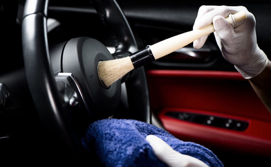 A gloved detailer using a soft detailing brush on the steering wheel and airbag of a car with a red leather interior.